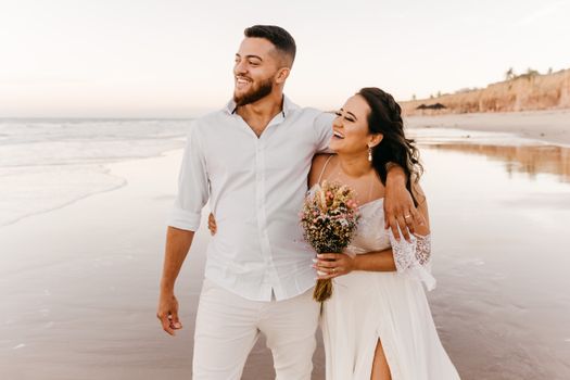 Smiling bearded groom in white shirt and trousers walking along sandy beach together with joyful bride in wedding dress