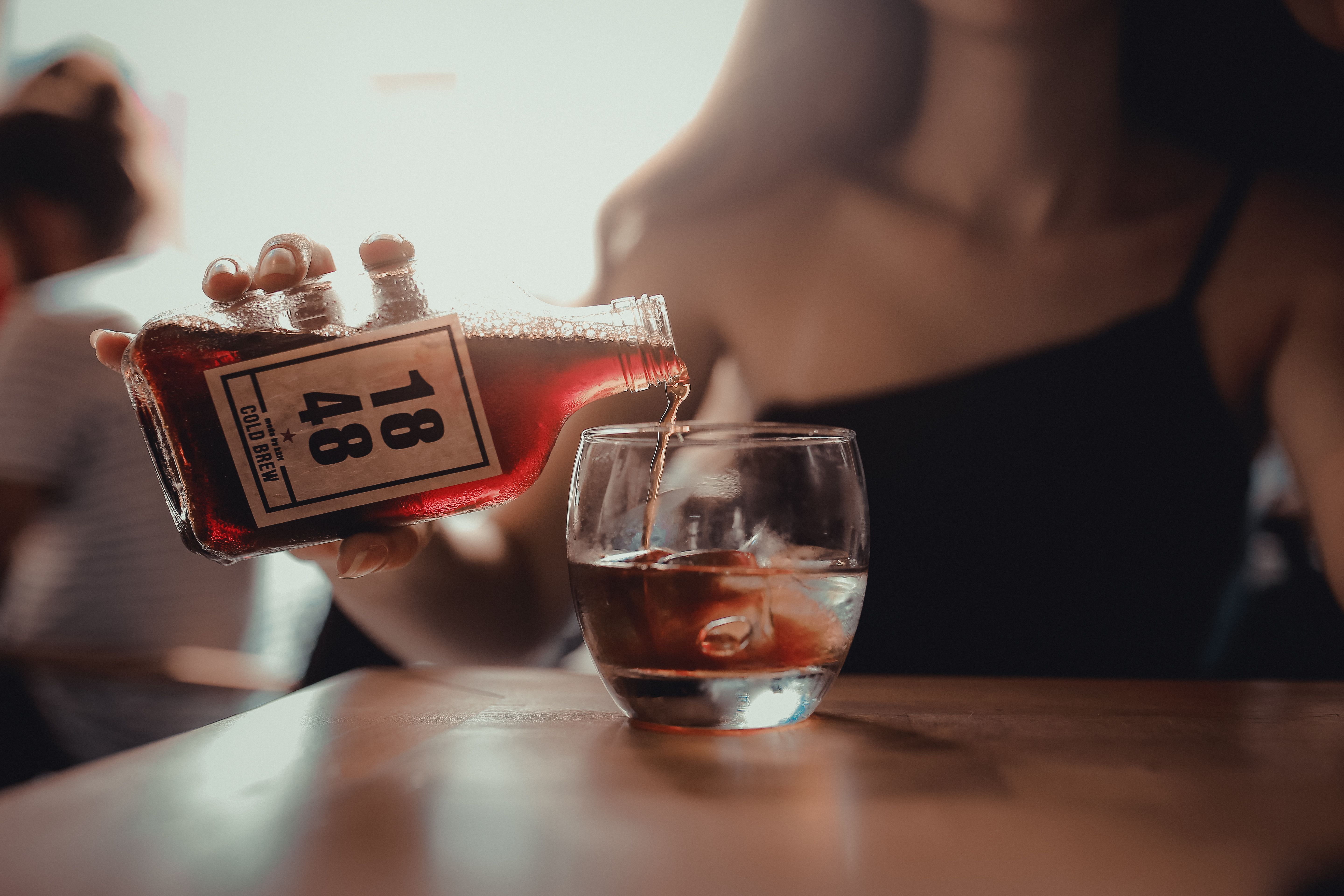 A woman pours cold brew coffee into a glass with ice cubes, featuring selective focus and warm tones.