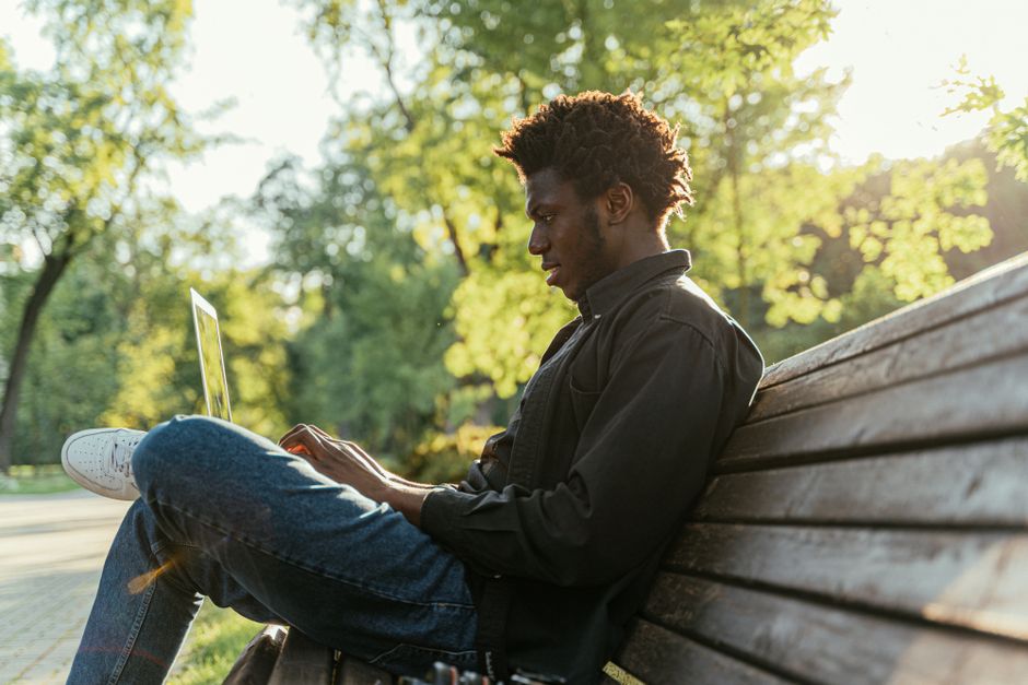 A young man sitting on a bench, focused on his laptop, surrounded by lush greenery. A young man sitting on a bench, focused on his laptop, surrounded by lush greenery.