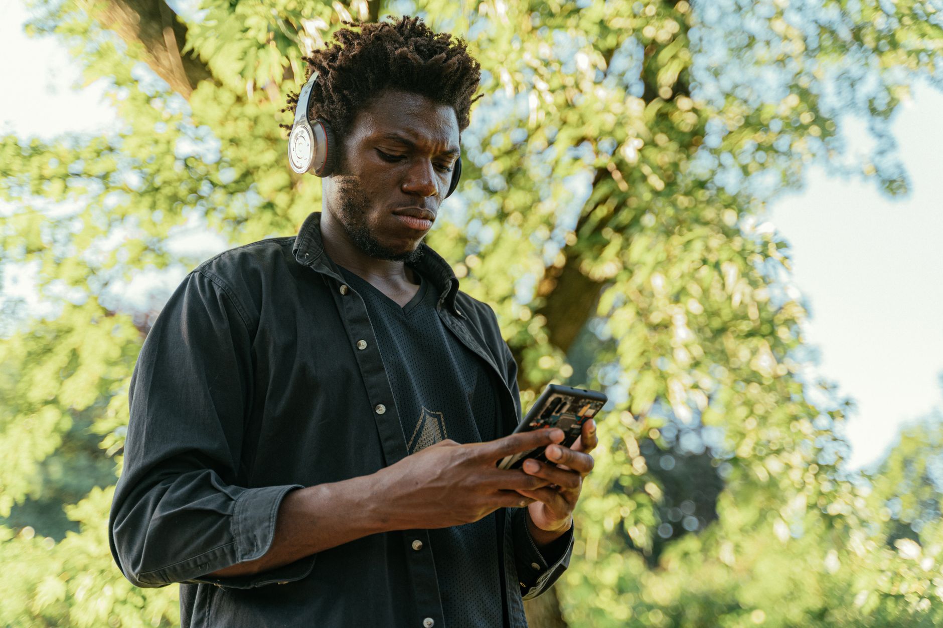 An adult man with headphones and afro hair using a smartphone in a tree-filled park setting.