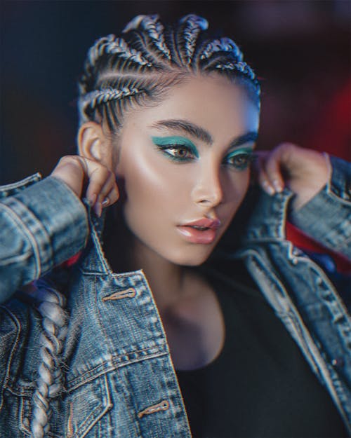 Young gorgeous female with vibrant makeup and cornrows looking down while adjusting collar of denim coat on blurred background