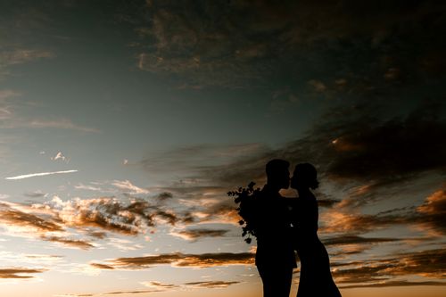 Free Silhouette of anonymous couple with flowers hugging and kissing in cloudy golden evening bright sky in darkness Stock Photo