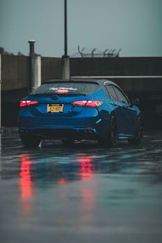 A sleek blue car on a rain-soaked road reflecting city lights, creating a moody atmosphere.