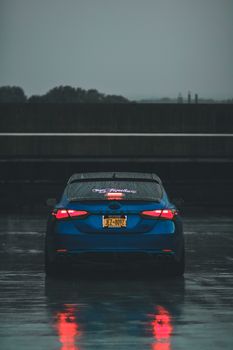 一辆时髦的蓝色汽车停在雨中的城市街道上，尾灯闪闪发光，映照着潮湿的人行道
