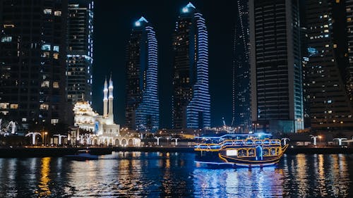 免費 白色和藍色的小船，在夜間城市建築物附近的水面上 圖庫相片