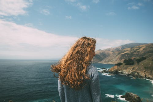 Free Woman Wearing Blue Long-sleeved Shirt Standing on Cliff Near Ocean Stock Photo