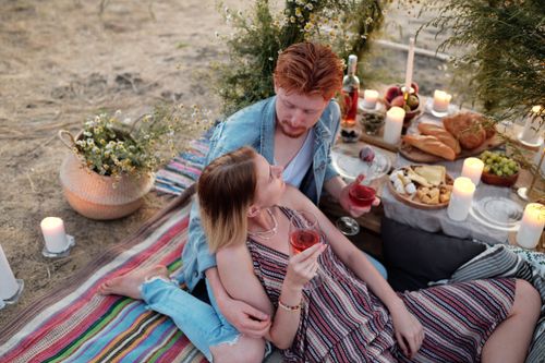 Free Couple enjoying a romantic picnic with wine, candles, and snacks outdoors at sunset. Stock Photo