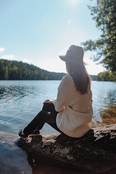 一名女子戴着太阳帽、穿着针织衫，安静地坐在宁静的湖边，享受着大自然