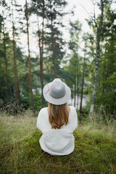 A woman in a knitted sweater sits in a grassy forest clearing, wearing a hat.