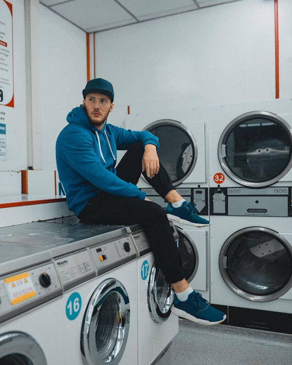 Man in Blue Long Sleeve Shirt and Black Pants Sitting on Front Load Washing Machine