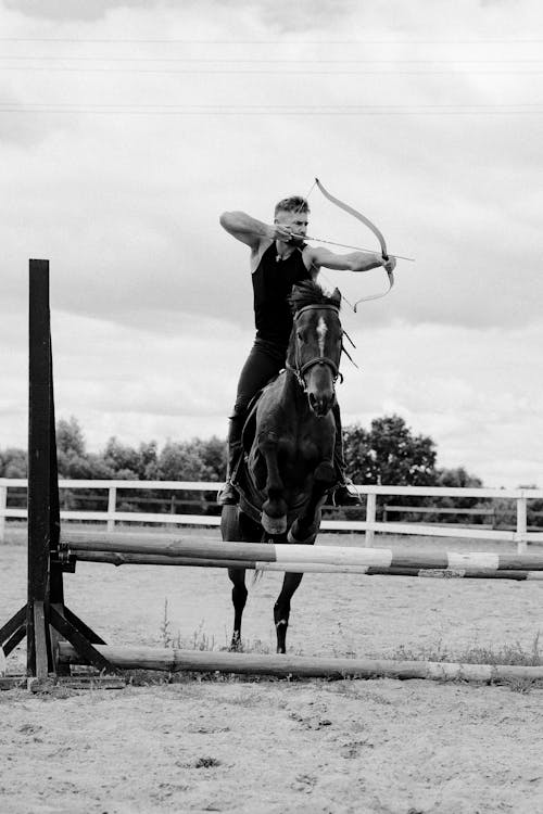 Grayscale Photo of Man Shooting an Arrow While Riding a Horse