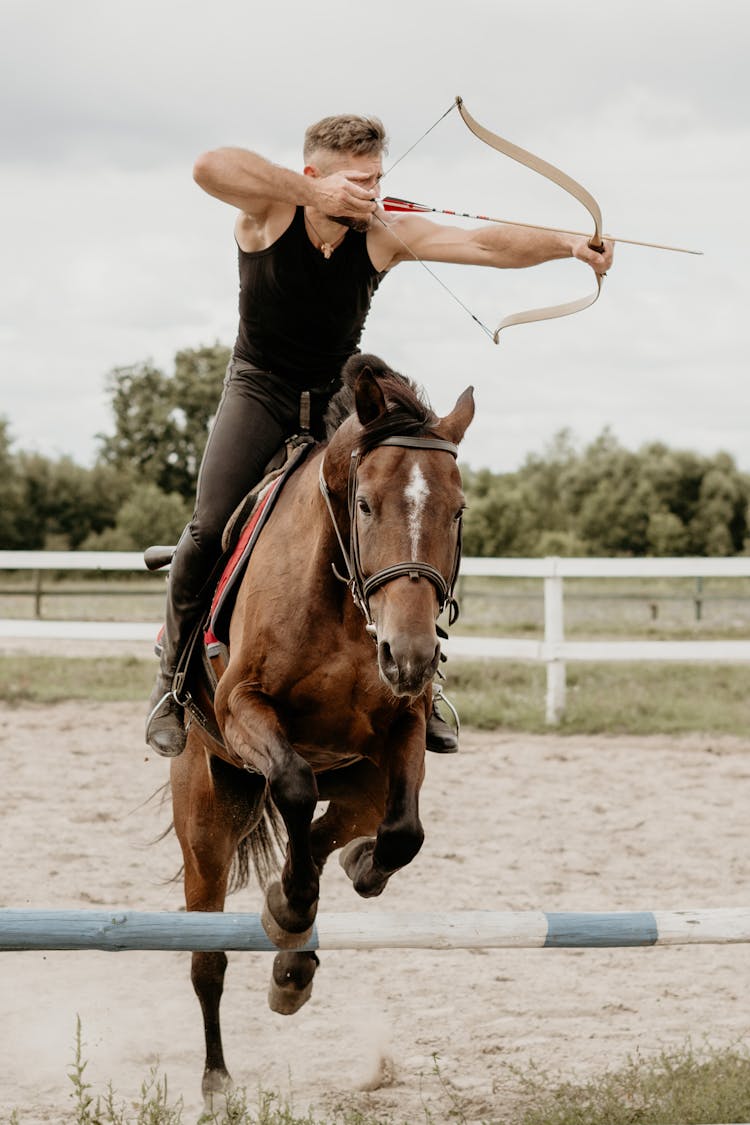 Man Shooting Bow From Horse Jumping Over Hurdle