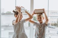 一对兄弟姐妹在窗前笑着，开玩笑地用头顶着书保持平衡，象征着学习的乐趣