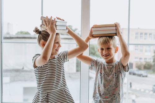Free Two laughing siblings playfully balance books on their heads by a window, symbolizing fun learning. Stock Photo