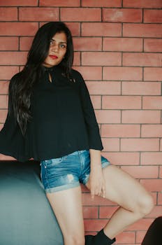 Serious ethnic female with red lips wearing denim shorts and black blouse standing against brick wall