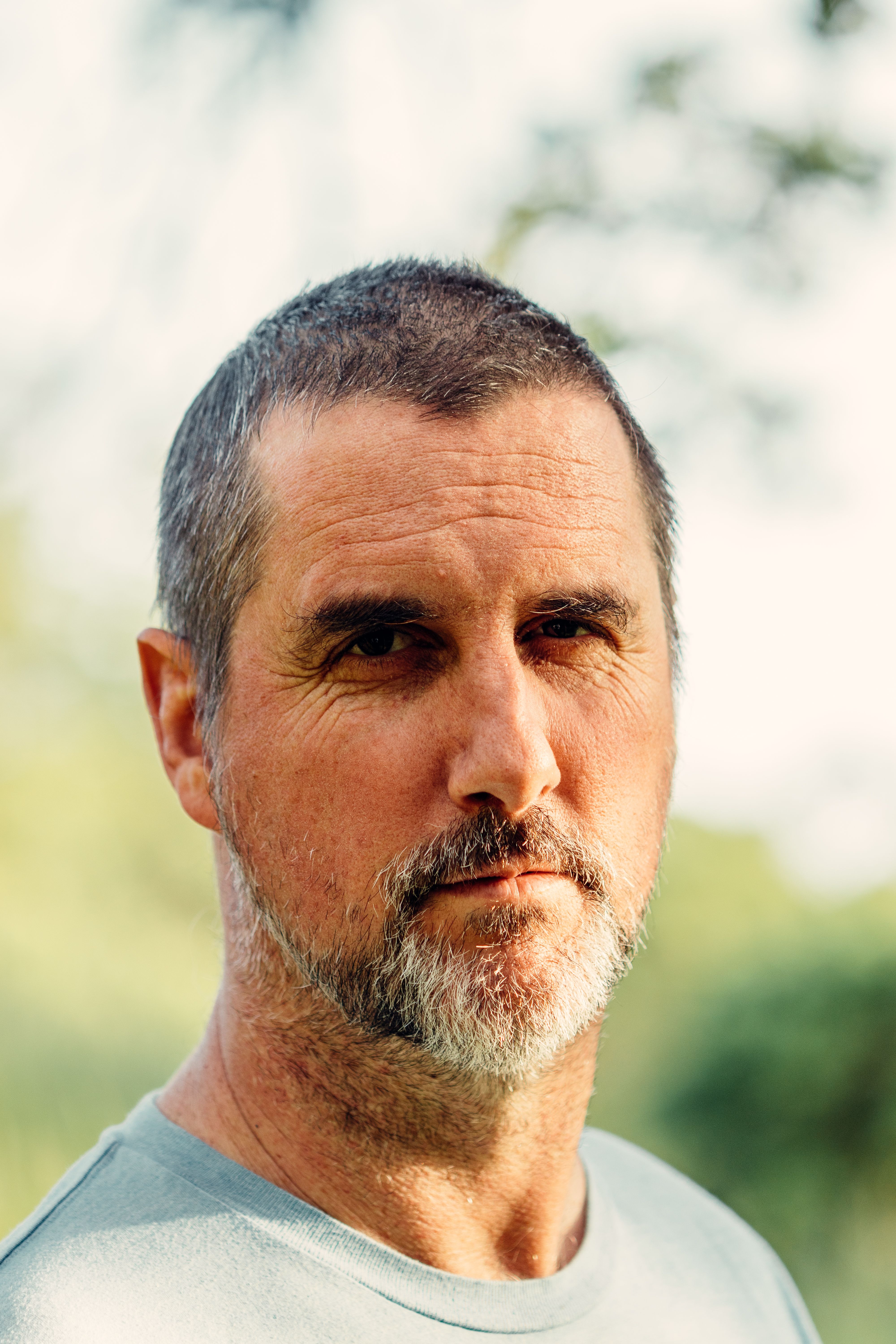 Bearded serious middle aged male in casual shirt standing against blurred lush trees and looking at camera calmly