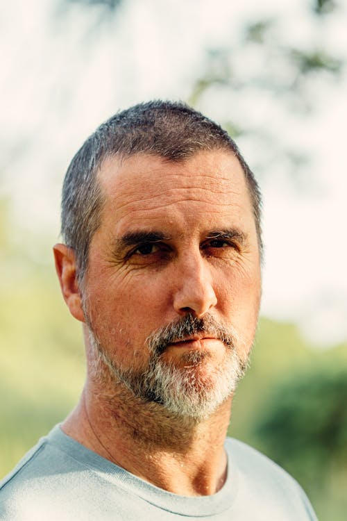 Free Bearded serious middle aged male in casual shirt standing against blurred lush trees and looking at camera calmly Stock Photo