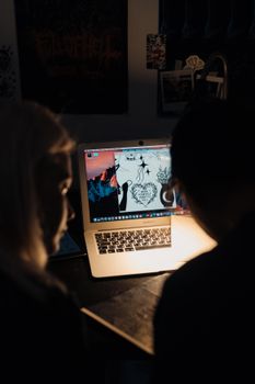 Two individuals observe digital art on a laptop screen in a dimly lit room.