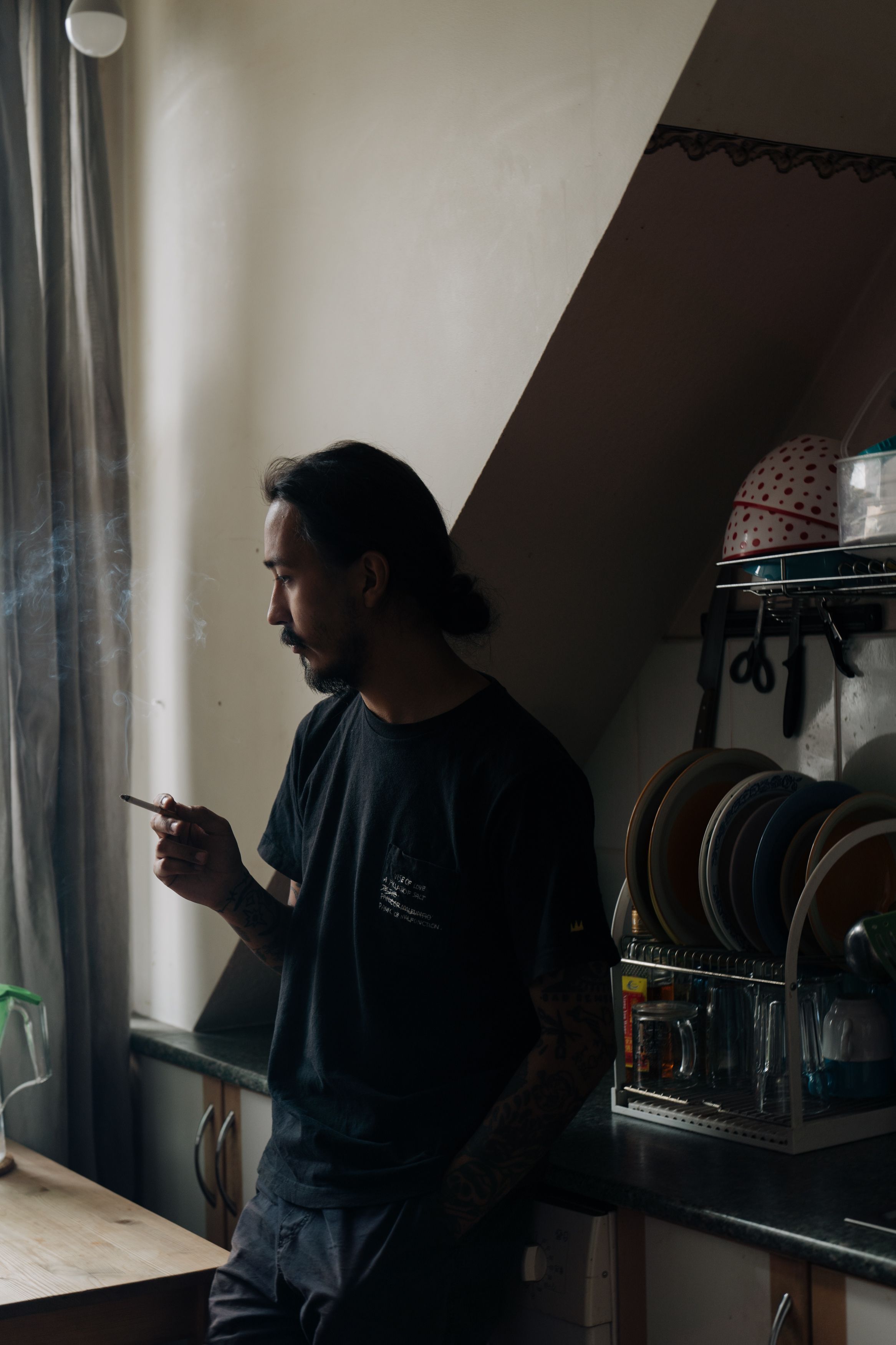 男人在柔和的自然光下靠近厨房窗户吸烟，陷入沉思