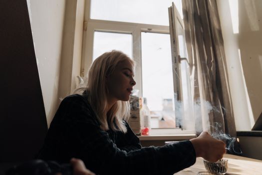 一位沉思的女人在敞开的窗边抽烟，阳光洒入，营造出一种忧郁的氛围