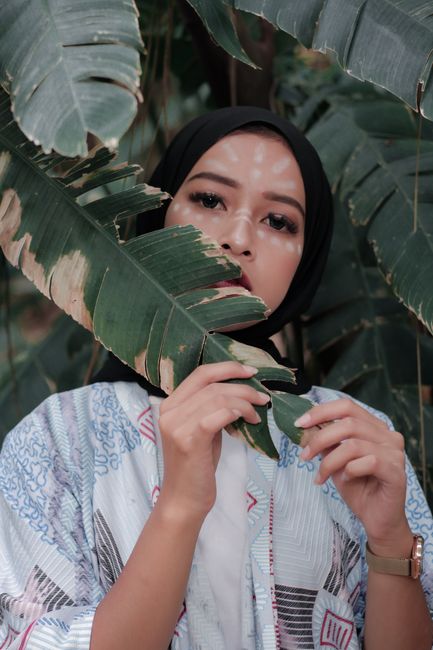 A woman in a hijab surrounded by lush, green tropical leaves, exhibiting grace and tradition.