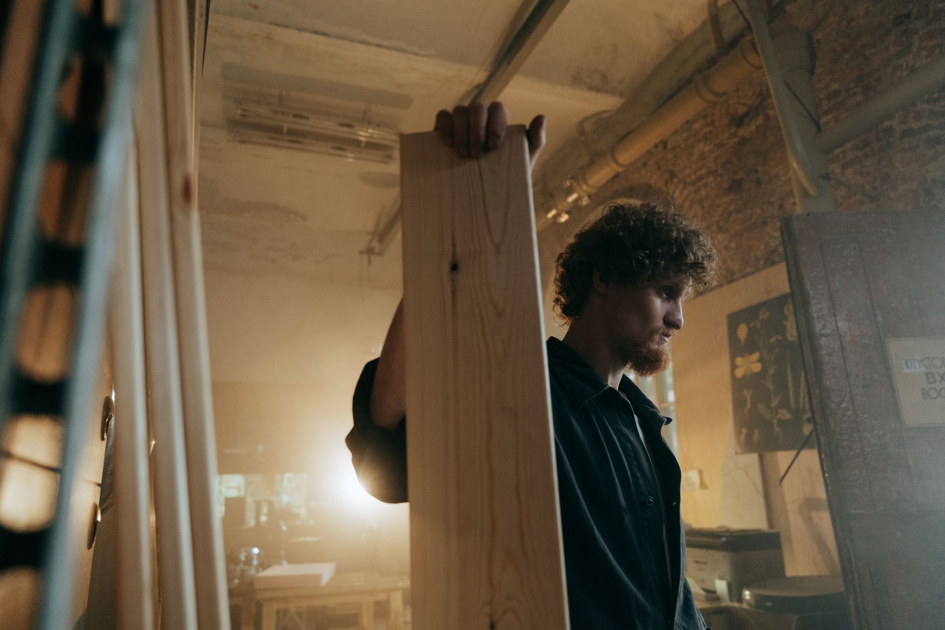 A male craftsman with curly hair works with wood in a rustic workshop setting.