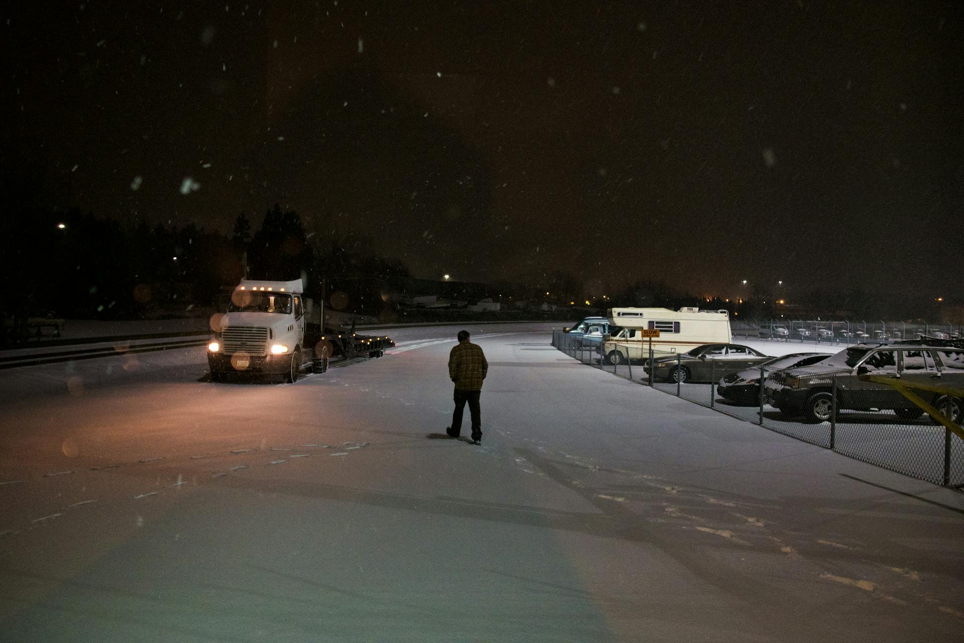 Lone figure and truck in snow-covered parking lot at night, lights in distance