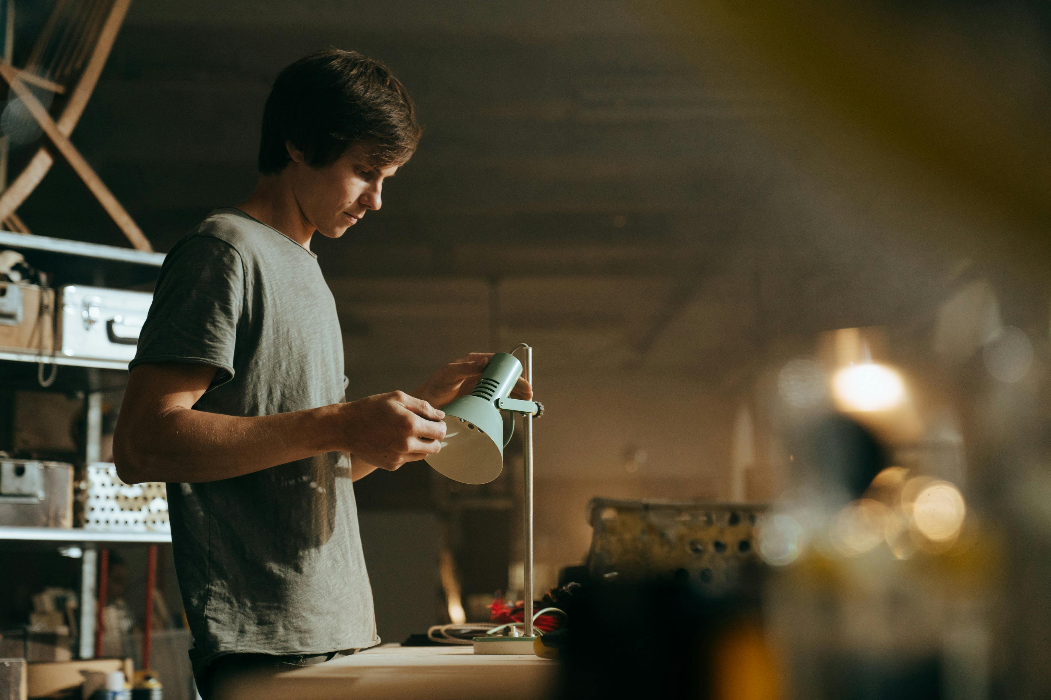 A young craftsman examines a lamp in his dimly lit workshop, focused and determined.