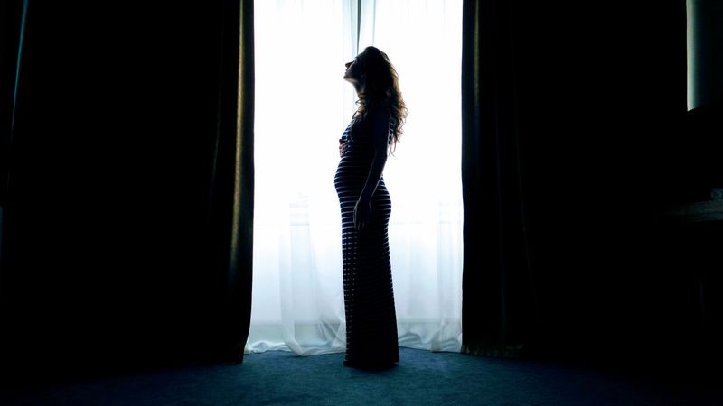 Silhouette of a pregnant woman standing by a bright window, captured in a serene indoor setting.