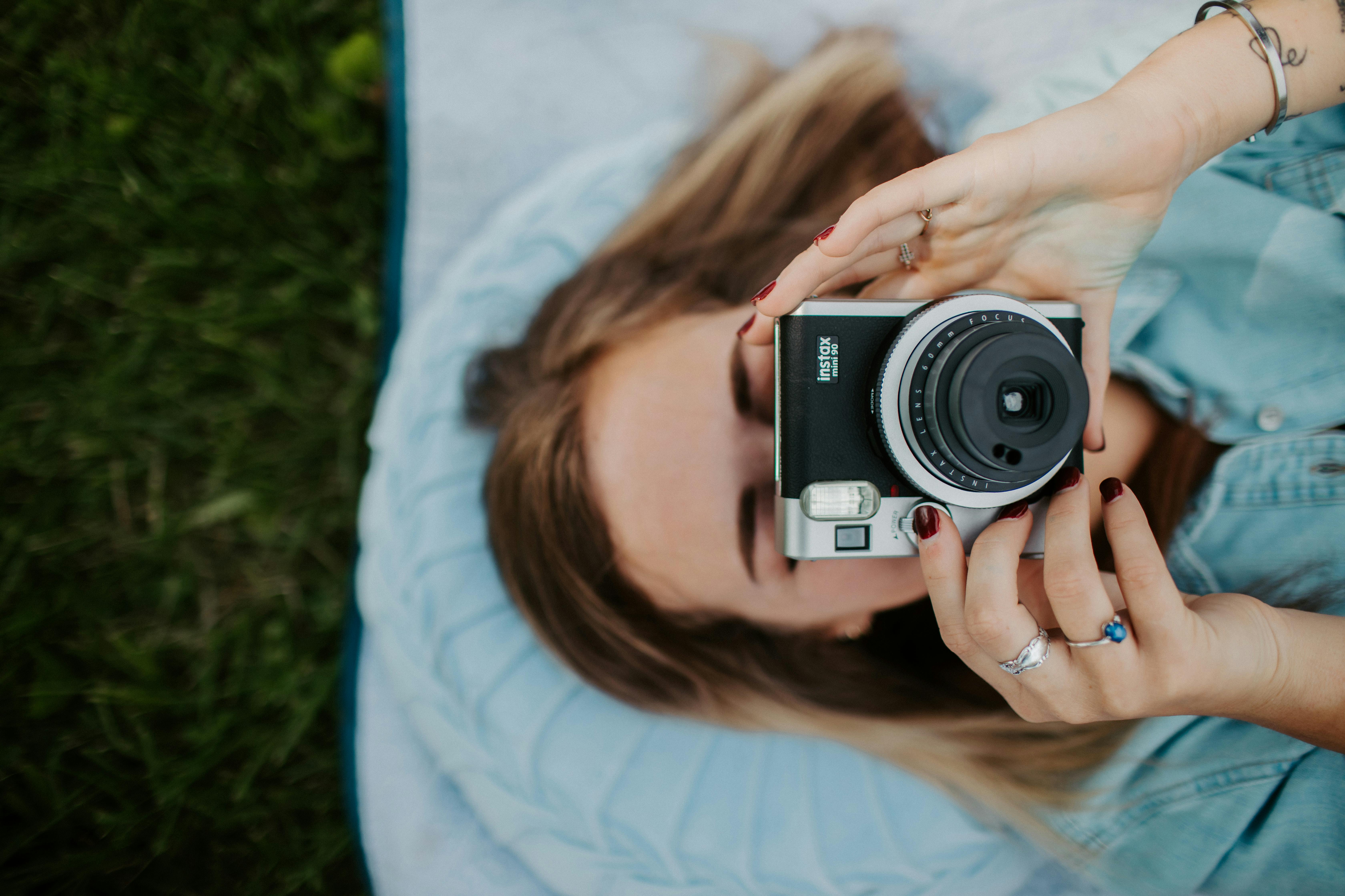 A woman lying on grass taking a photo with an instant camera, captured from above.