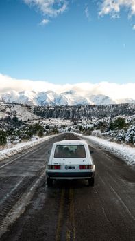 A vintage car travels on a snowy highway in Argentina, with majestic mountains in the backdrop.