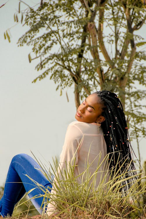From below back view of young mindful African American lady in trendy outfit resting on meadow with closed eyes under sky