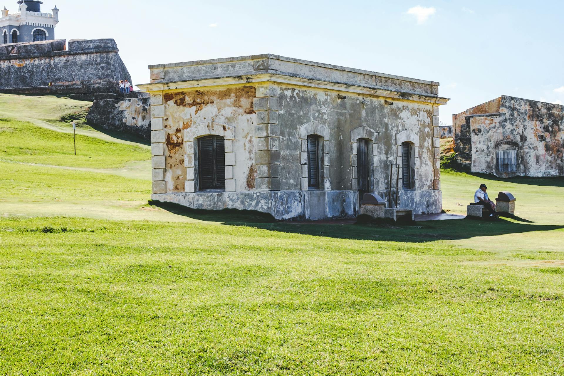 A historic stone building located on a green hill with an old fort in the background, under a clear sky.