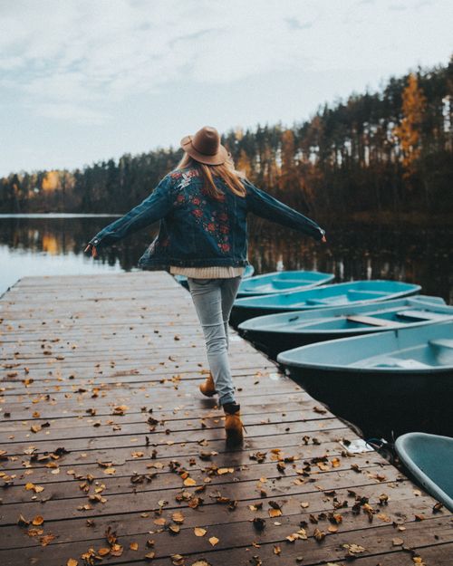 Free A woman in a denim jacket walks along a wooden dock beside a serene lake with rowboats. Stock Photo