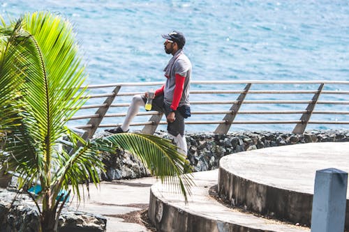 Free A Man in Gray Shirt and Black Cap Holding a Plastic Bottle of Drink while Looking at the Calm Water of the Sea Stock Photo