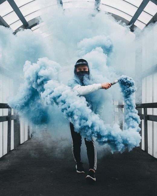 A person wearing a cap and facemask holds a blue smoke bomb inside a modern enclosed walkway.