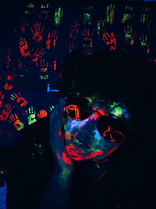 Serious male in sunglasses with colorful glowing neon paint on face in dark room with Uv lights