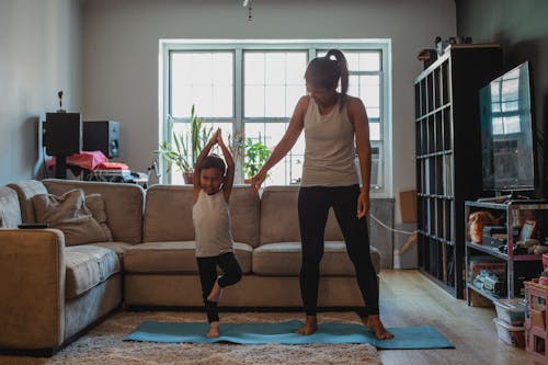 Full body positive mother teaching little kid to perform Tree Pose on mat in cozy living room at home