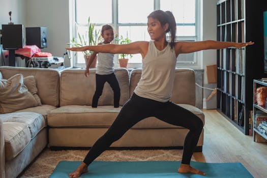Full body barefoot cheerful female in sportswear standing in Warrior Pose on mat in living room near cute daughter walking on sofa