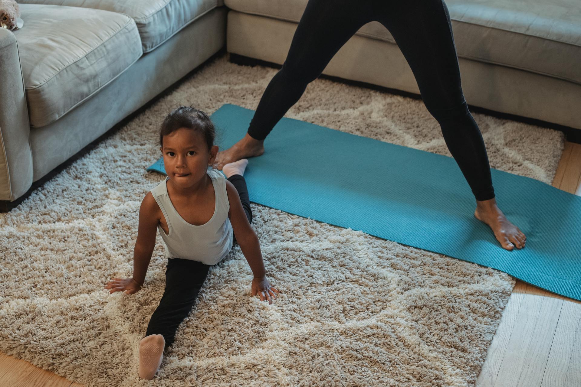 High angle flexible ethnic girl in sportswear doing splits on fluffy carpet near mother while training together at home