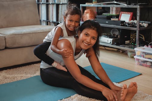 Free Happy ethnic mother stretching body and piggybacking adorable daughter while sitting on mat at home and looking at camera Stock Photo