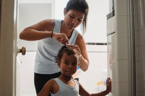 Young ethnic mother doing hair of cute daughter in bathroom