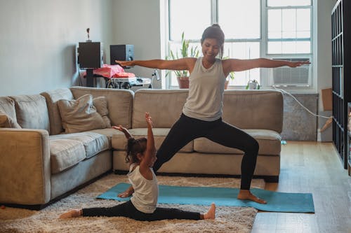 Full body cheerful ethnic mother in sportswear practicing yoga and performing Warrior Pose on mat near cute daughter doing splits in light living room