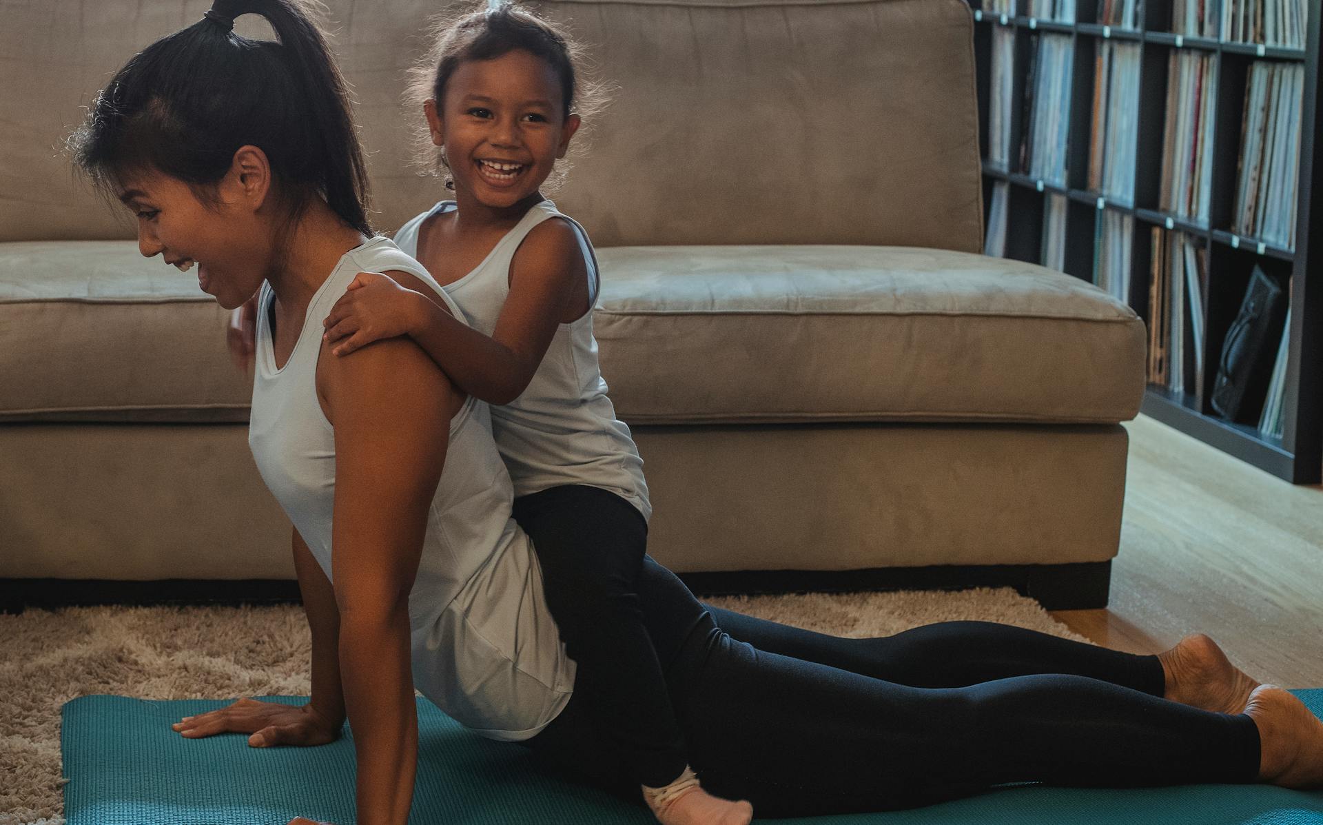 Side view laughing young ethnic female in sportswear performing Cobra Pose with cute smiling daughter on back while practicing yoga at home