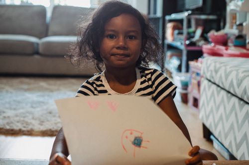 Free Cute ethnic girl in casual shirt demonstrating paper sheet with drawing while sitting in living room and looking at camera Stock Photo