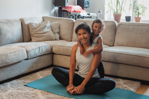 Full body happy ethnic girl hugging smiling young mother sitting on mat in Lotus Pose at home while looking at camera