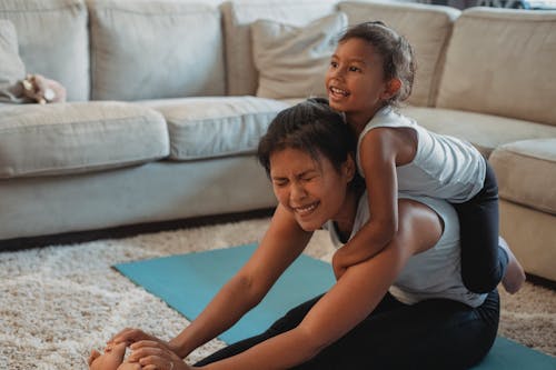 Cheerful small Hispanic daughter riding piggyback on mother stretching on mat in living room in daytime