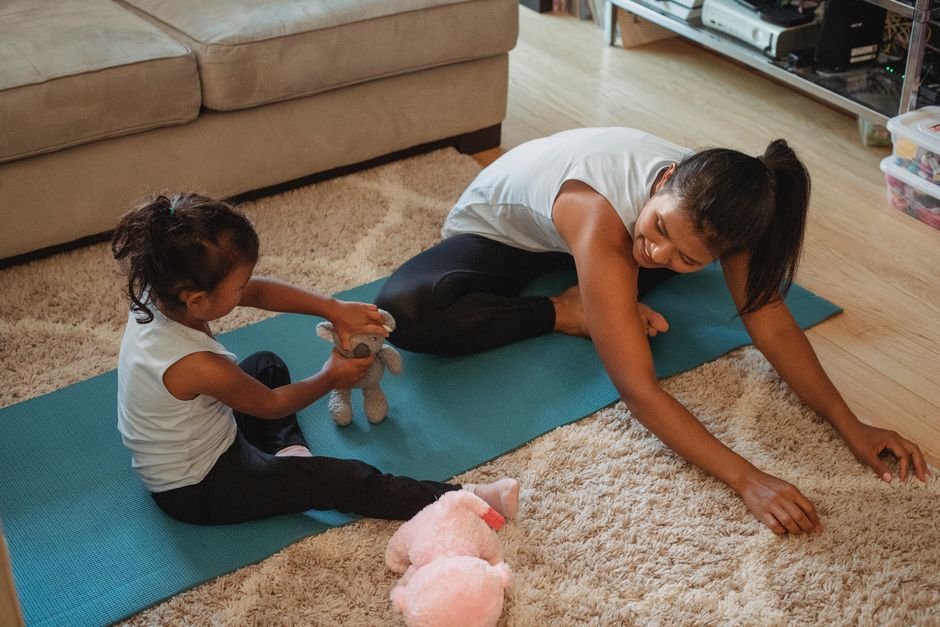 Primary keyword — A tired but smiling Indian working mom doing a simple stretching exercise in her living room while a child plays nearby on a colorful mat