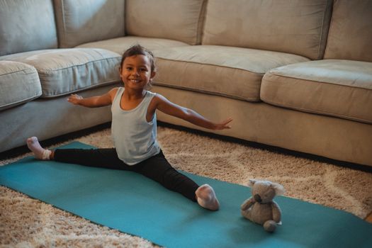 Cheerful little ethnic little girl stretching on stretch mat at home with toy and looking at camera