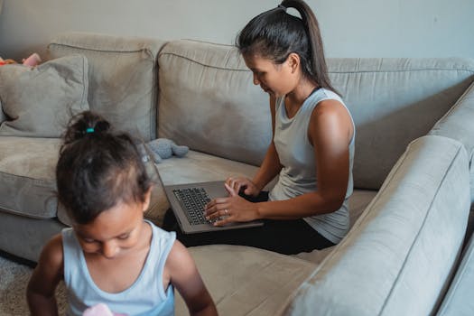 Adult ethnic woman using laptop while sitting on sofa with little daughter playing with toys at home